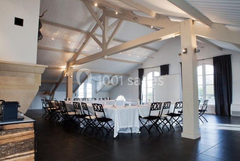 Salle lumineuse avec grande table rectangulaire dressée pour une réunion, entourée de chaises noires, sous une charpente…