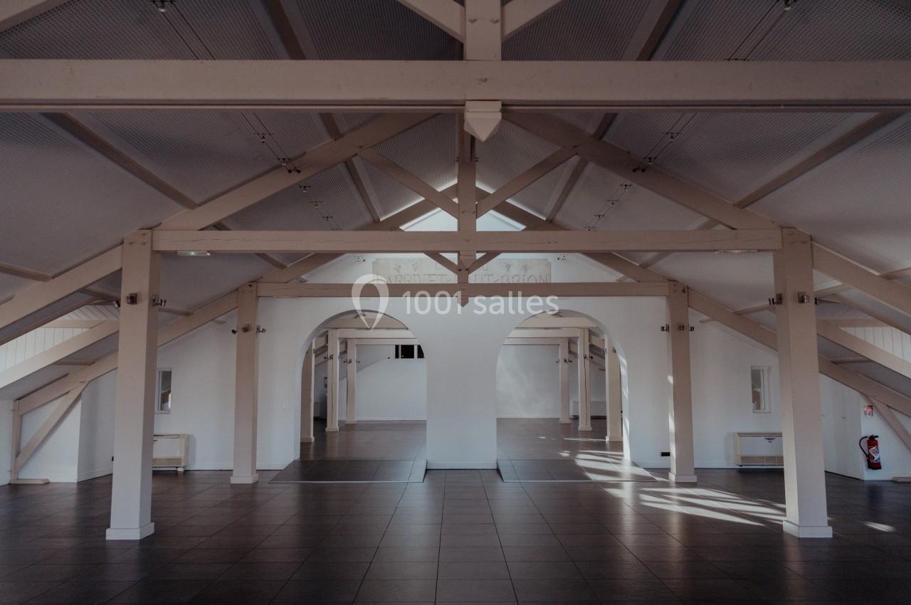 Intérieur d'un grand espace lumineux avec charpente apparente, murs blancs et sol en carrelage sombre.