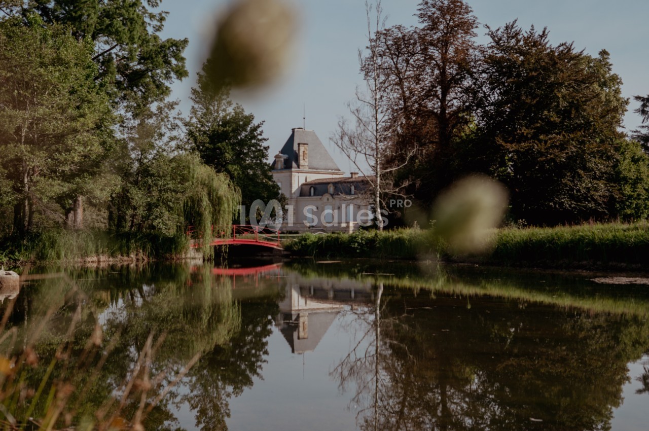 Vue d'un château entouré de végétation, reflété dans un étang avec un pont rouge au premier plan.