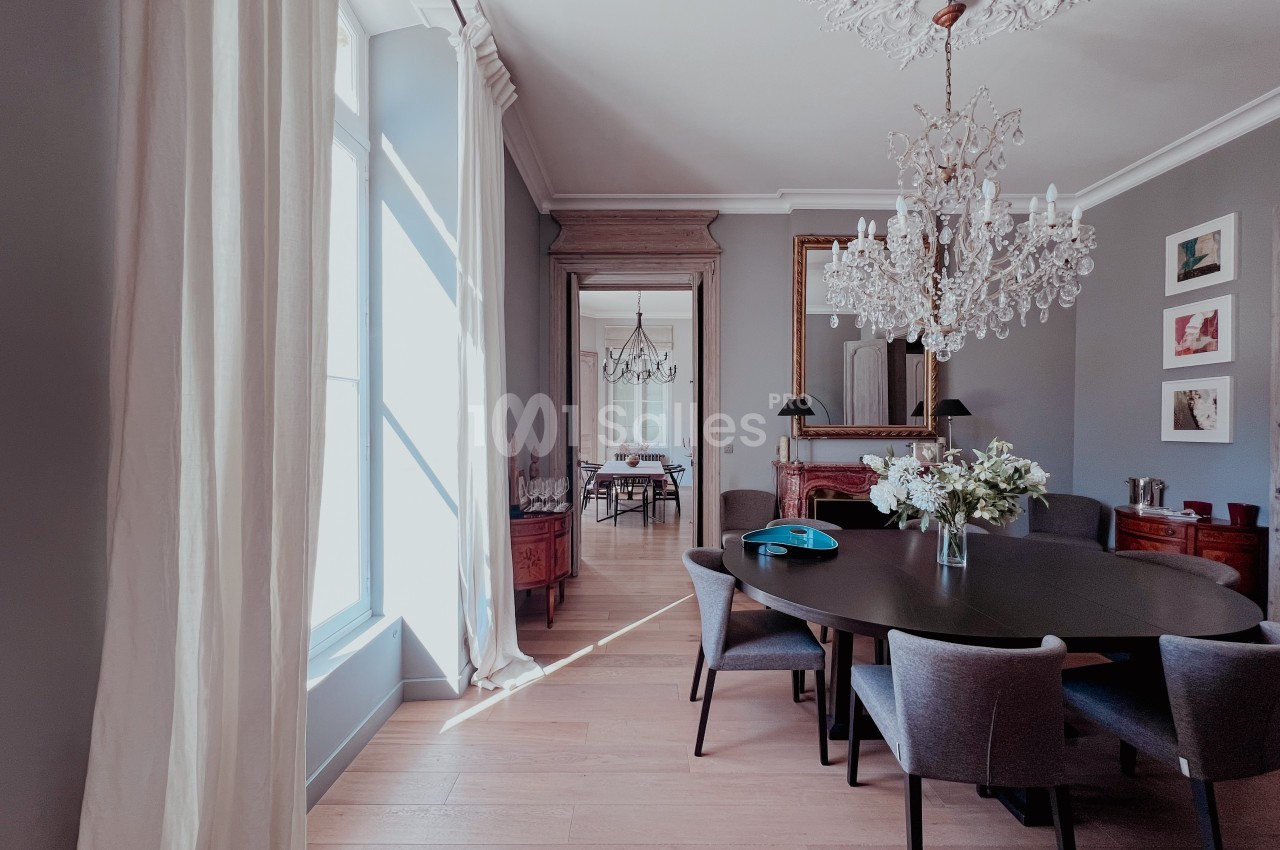 Salle à manger élégante avec table en bois, chaises grises, lustre en cristal et grande fenêtre laissant entrer la lumière.