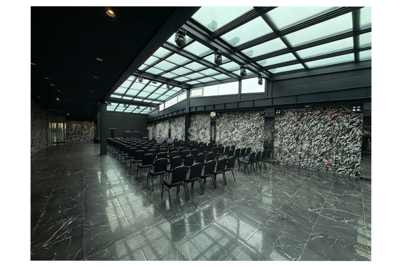 Salle de conférence moderne avec des rangées de chaises noires, sol en marbre et toit vitré laissant entrer la lumière…