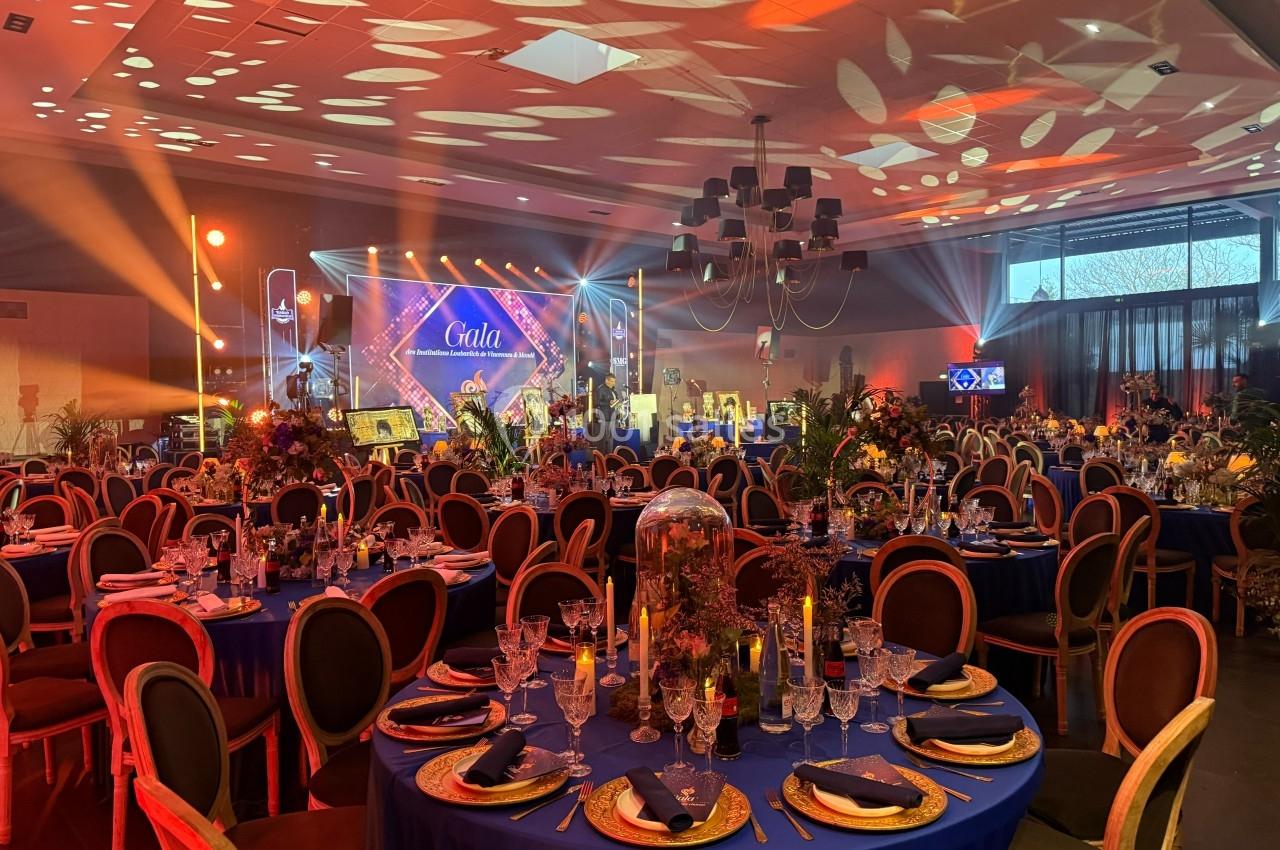Salle de réception décorée pour un gala, avec tables dressées, éclairages colorés et scène en arrière-plan.