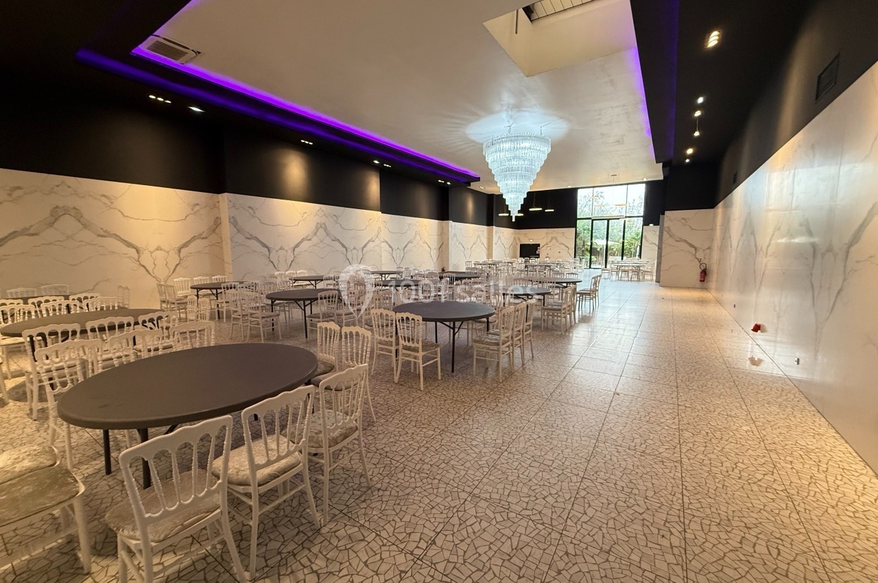 Salle de réception spacieuse avec tables rondes, chaises blanches, murs effet marbre, lustre central et éclairage violet.