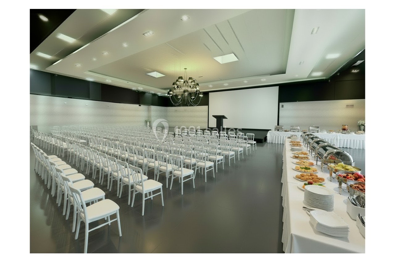 Salle de conférence avec rangées de chaises blanches, un podium, un écran de projection et un buffet dressé sur le côté.
