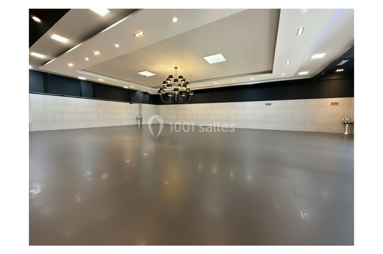 Salle spacieuse et vide avec un sol gris, un grand lustre central et des murs clairs ornés de panneaux.