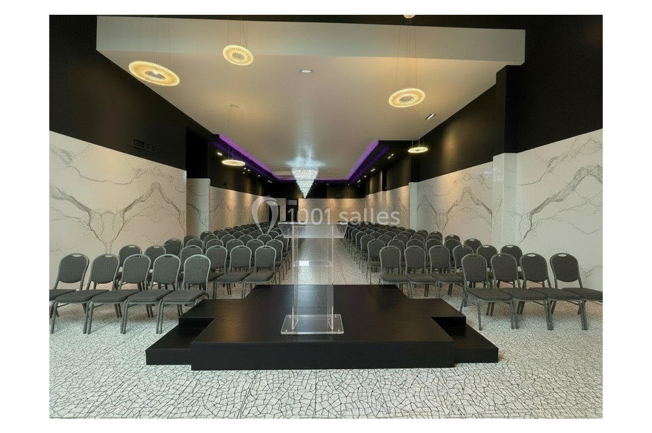 Salle de conférence moderne avec rangées de chaises, estrade centrale et éclairage suspendu élégant.