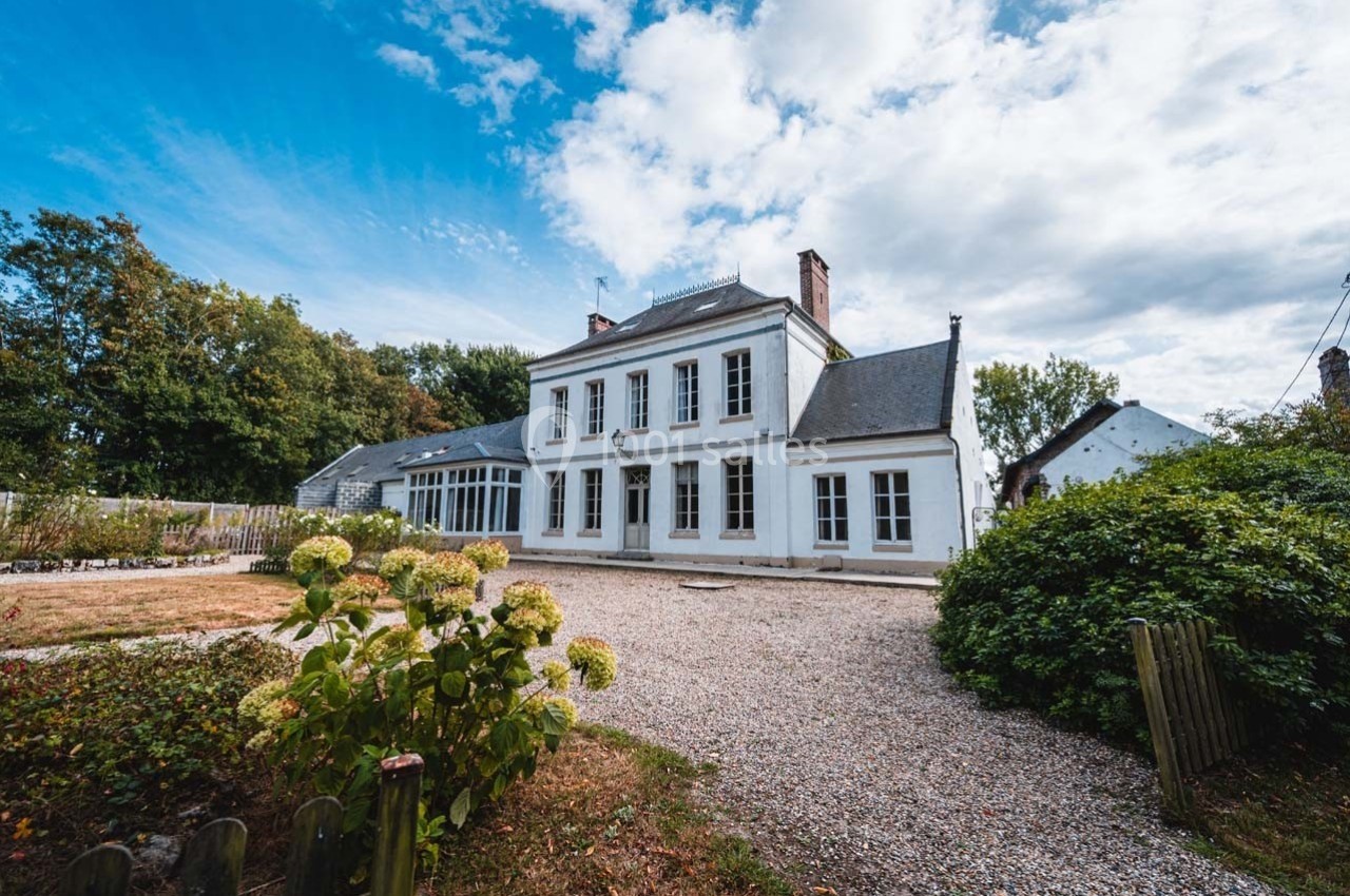 Façade d'une grande maison blanche avec jardin, allée gravillonnée et ciel partiellement nuageux en arrière-plan.