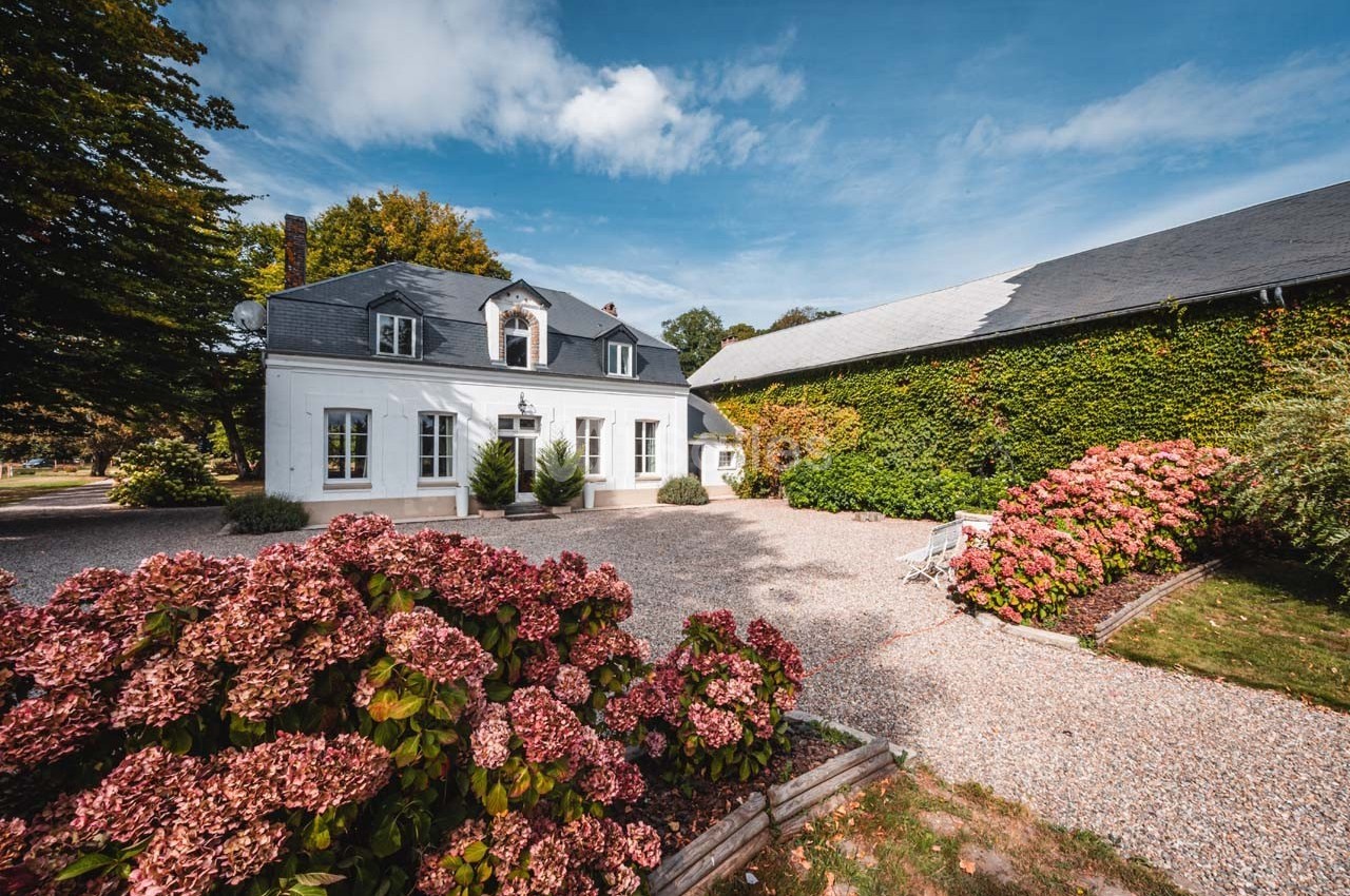 Maison blanche avec toit en ardoise, entourée d'hortensias et d'une cour gravillonnée, sous un ciel bleu.
