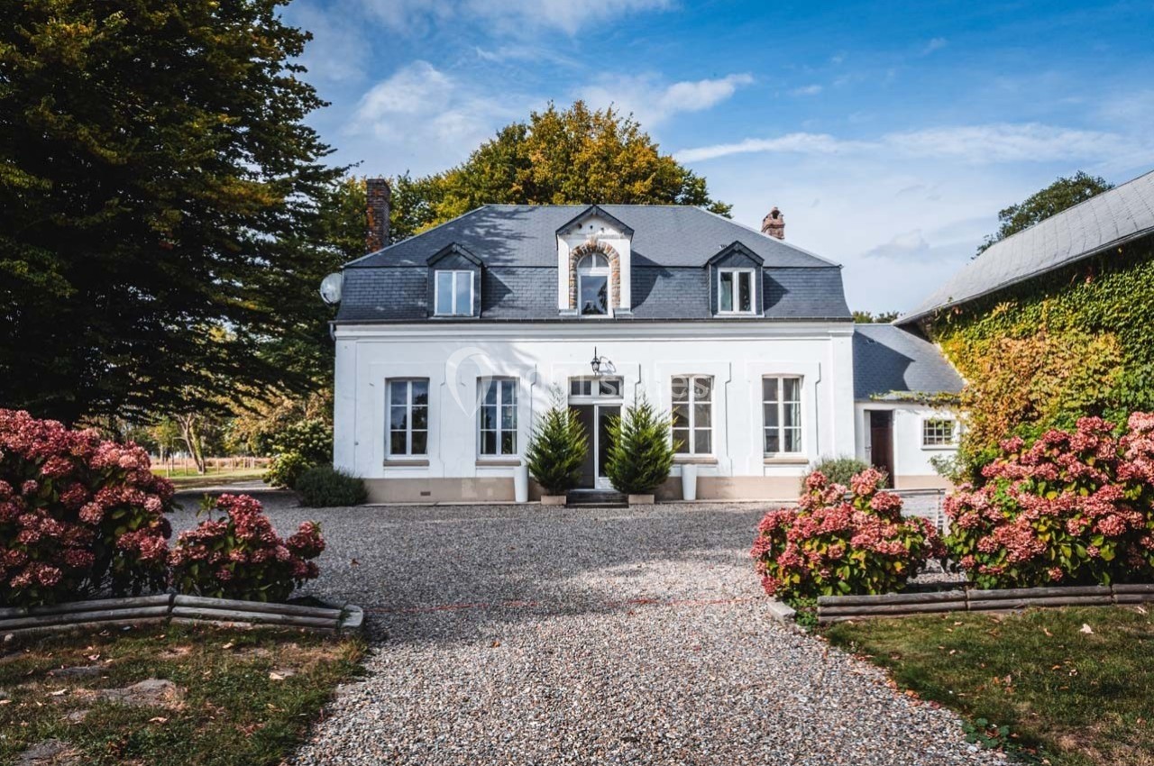 Façade d'une maison blanche avec toit en ardoise, entourée d'arbustes fleuris et d'une allée en gravier.