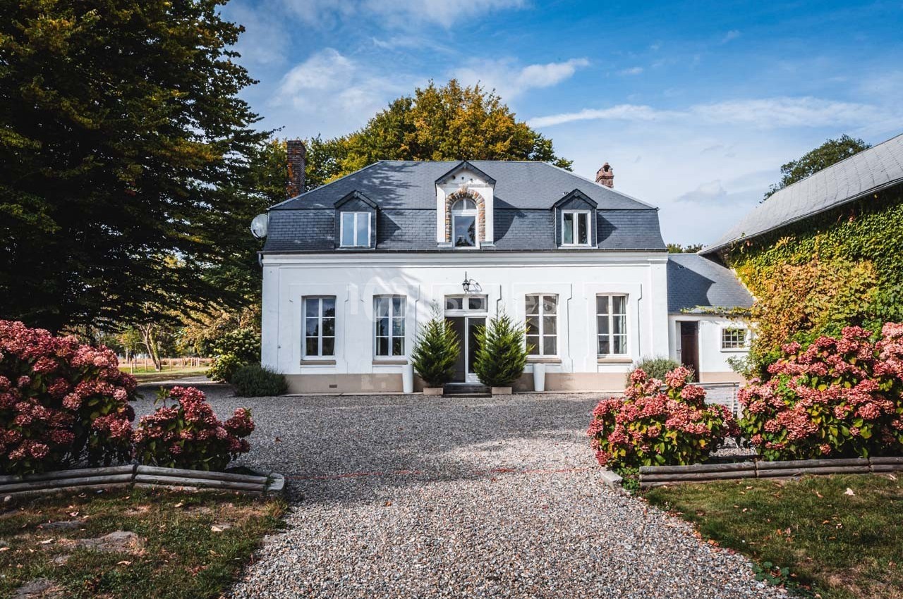 Façade d'une maison blanche avec toit en ardoise, entourée d'arbustes fleuris et d'une allée en gravier.