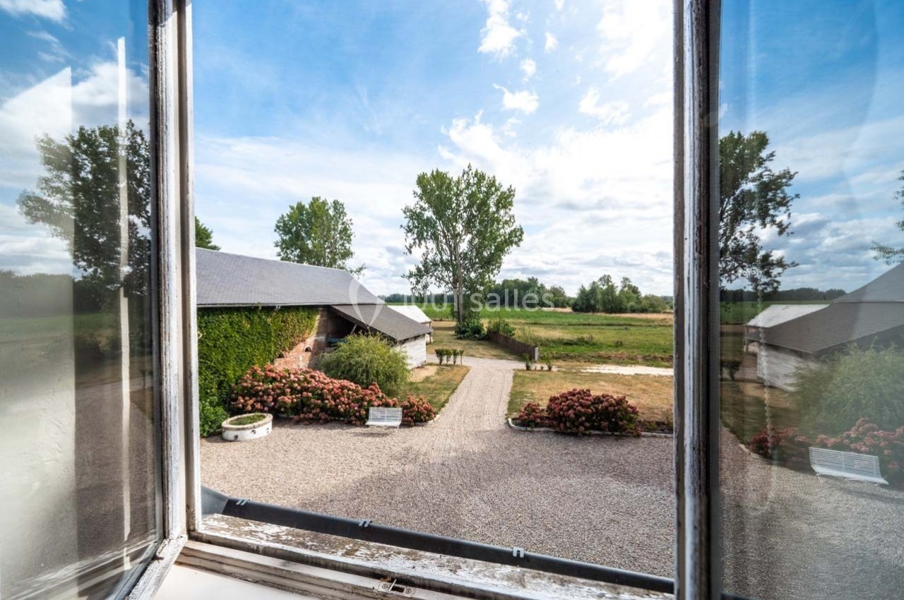 Vue depuis une fenêtre ouverte sur une cour gravillonnée, entourée de bâtiments et de végétation, sous un ciel partiellement…