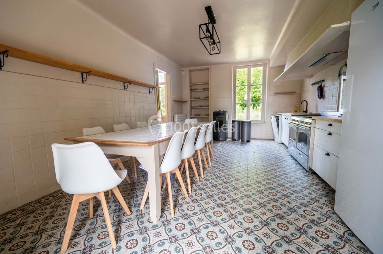Grande cuisine lumineuse avec table en bois, chaises blanches, carrelage à motifs et vue sur un jardin.