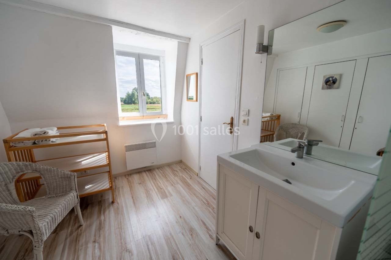 Salle de bain lumineuse avec lavabo, meuble blanc, chaise en osier, table à langer en bois et fenêtre donnant sur l…