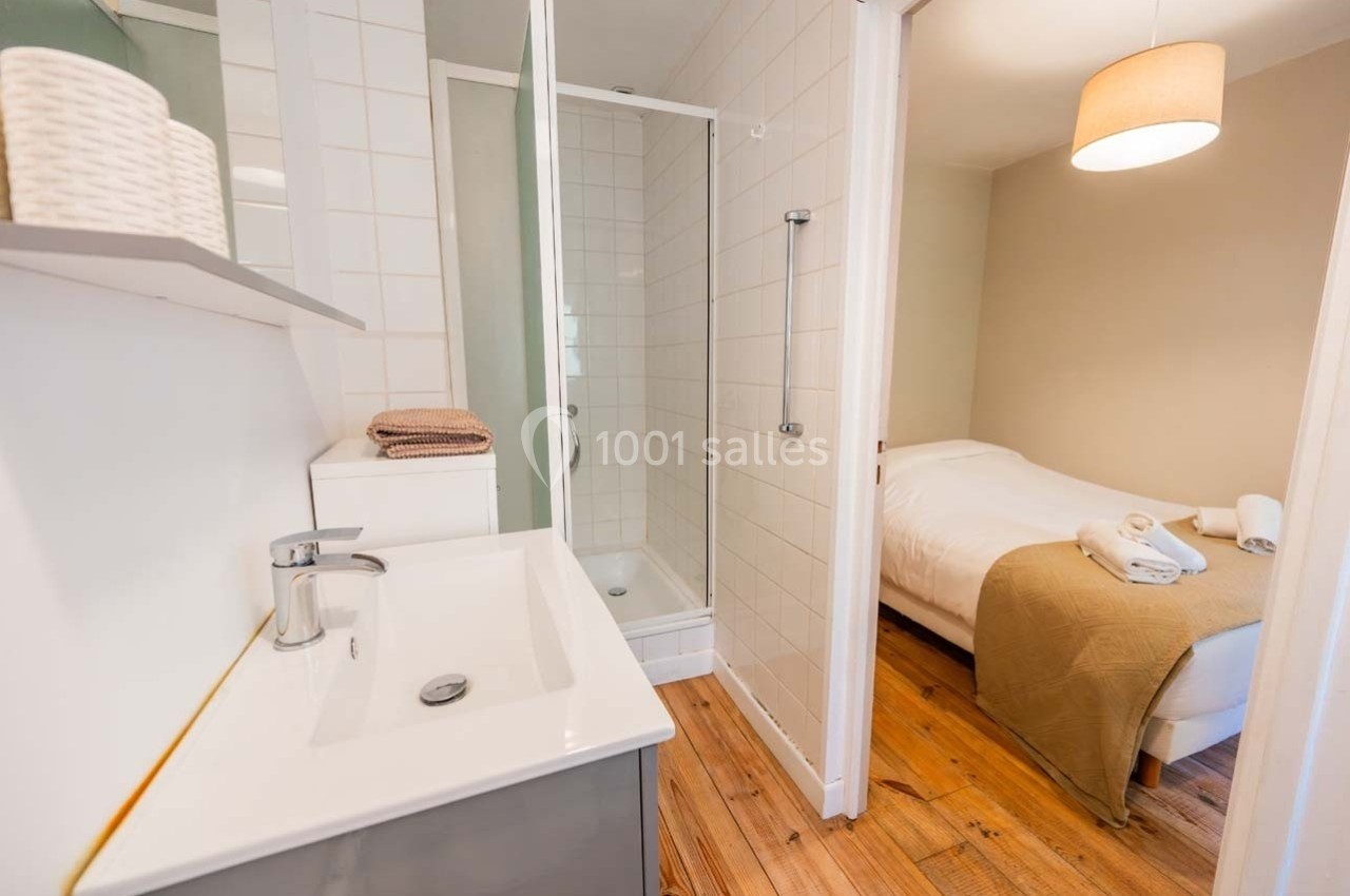 Salle de bain avec lavabo, douche et vue sur une chambre équipée d'un lit double avec linge de lit beige.