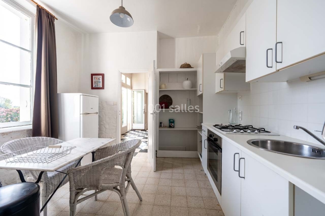 Cuisine lumineuse avec mobilier blanc, table en osier, réfrigérateur, et fenêtre donnant sur l'extérieur.