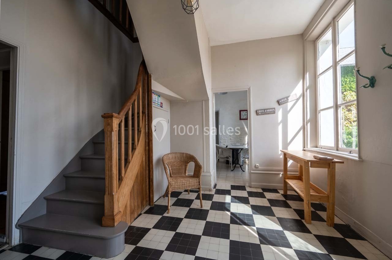 Entrée lumineuse avec escalier en bois, sol en carreaux noirs et blancs, chaise en osier et table près d'une fenêtre.
