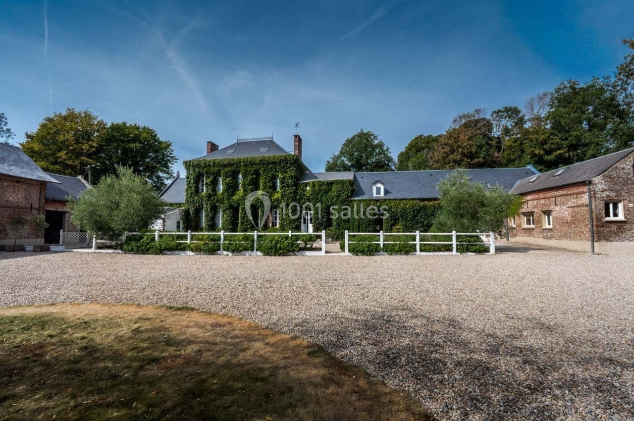 Grande maison en pierre recouverte de lierre, entourée d'arbres, avec une cour en gravier au premier plan.