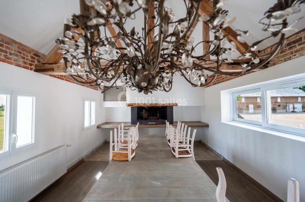 Salle lumineuse avec poutres apparentes, lustre décoratif, cheminée centrale et chaises en bois autour de tables.