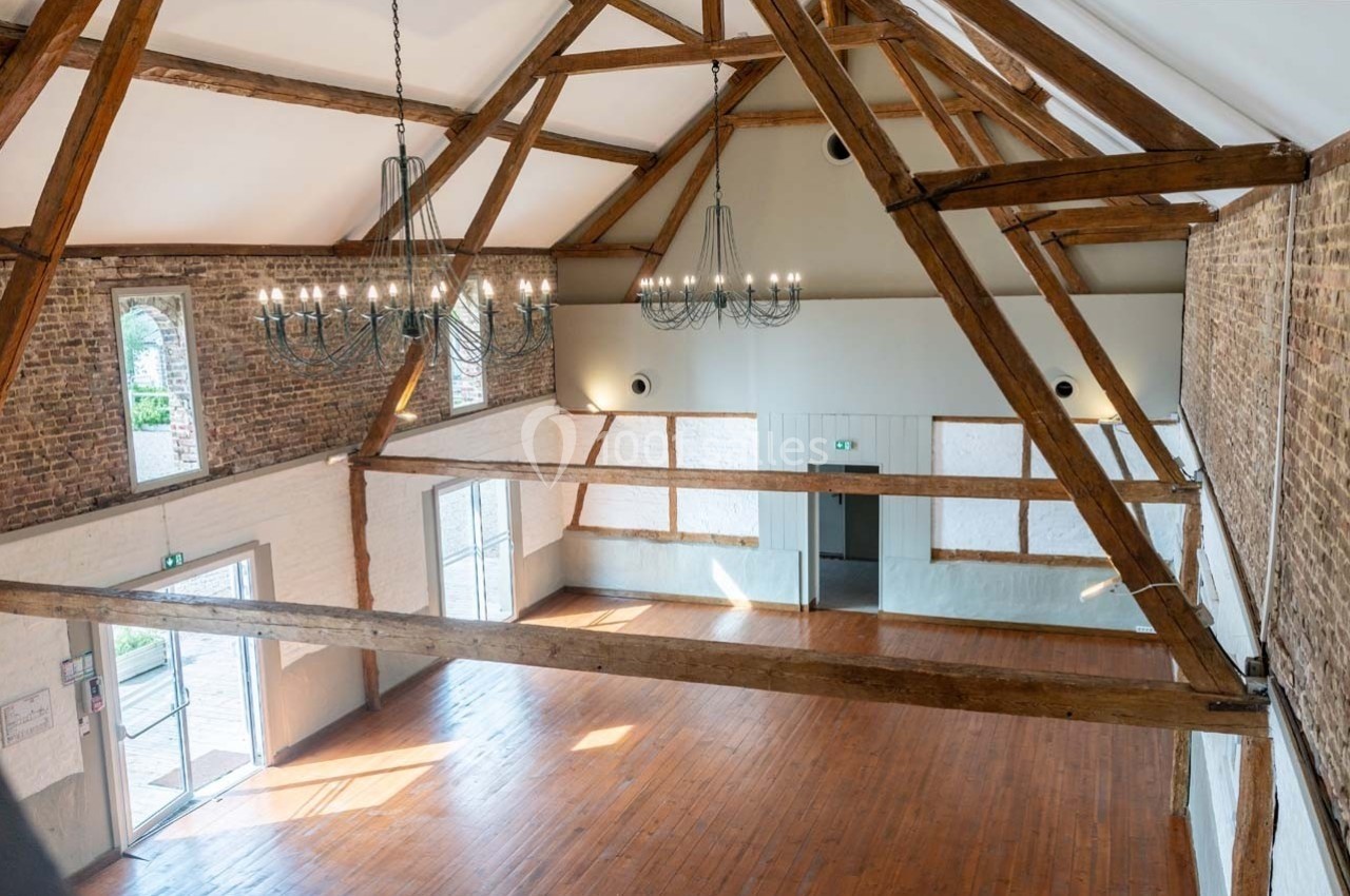 Salle lumineuse avec poutres apparentes, murs en briques et parquet, éclairée par des lustres et des fenêtres.