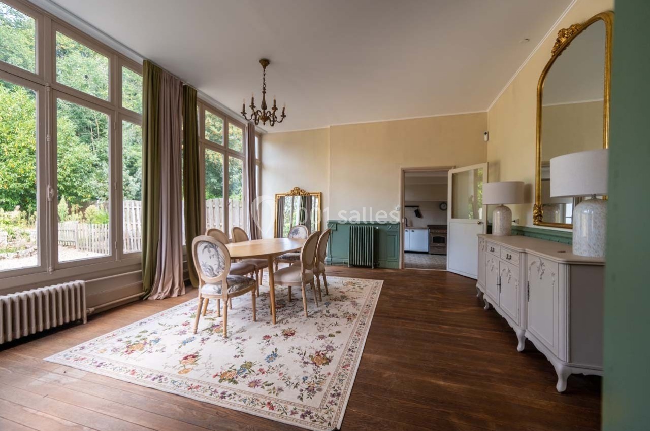 Salle à manger lumineuse avec table en bois, chaises élégantes, tapis fleuri, miroir doré et grandes fenêtres donnant sur un…