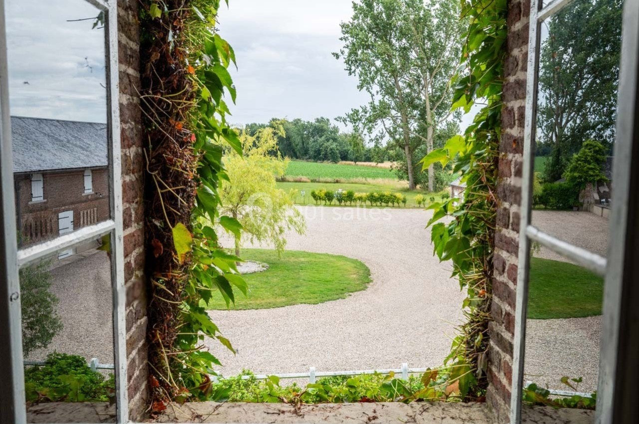 Vue depuis une fenêtre encadrée de lierre, donnant sur une cour gravillonnée et un paysage verdoyant.