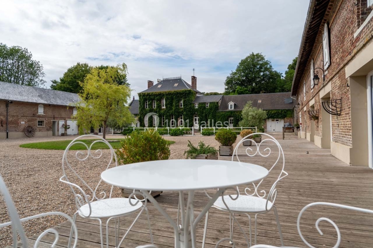 Table ronde blanche et chaises en fer forgé sur une terrasse en bois, avec vue sur une grande bâtisse couverte de lierre.