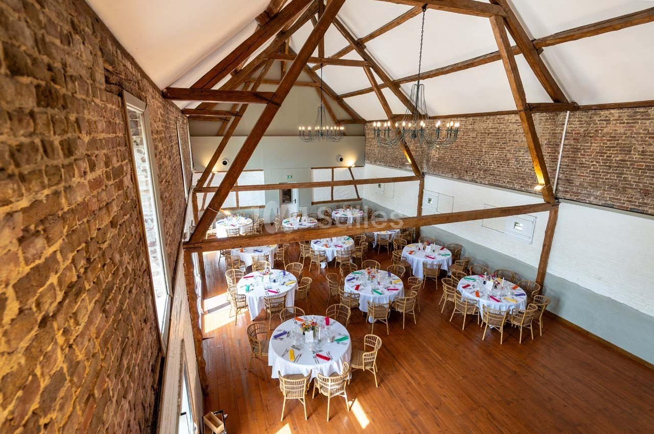 Salle de réception avec murs en briques, poutres apparentes et tables rondes dressées pour un événement.