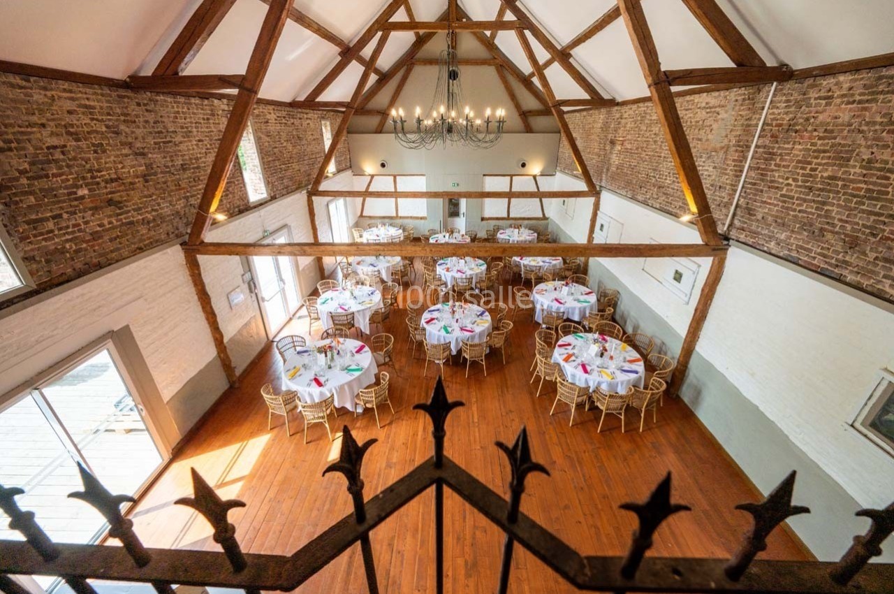 Salle de réception avec charpente en bois apparente, tables rondes dressées et sol en parquet.