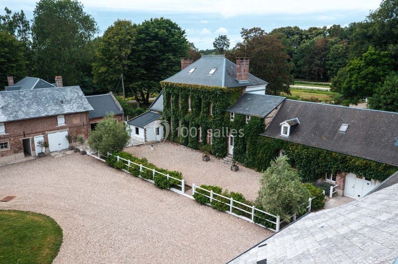 Vue aérienne d'une grande maison en briques recouverte de lierre, entourée d'une cour gravillonnée et de bâtiments annexes.