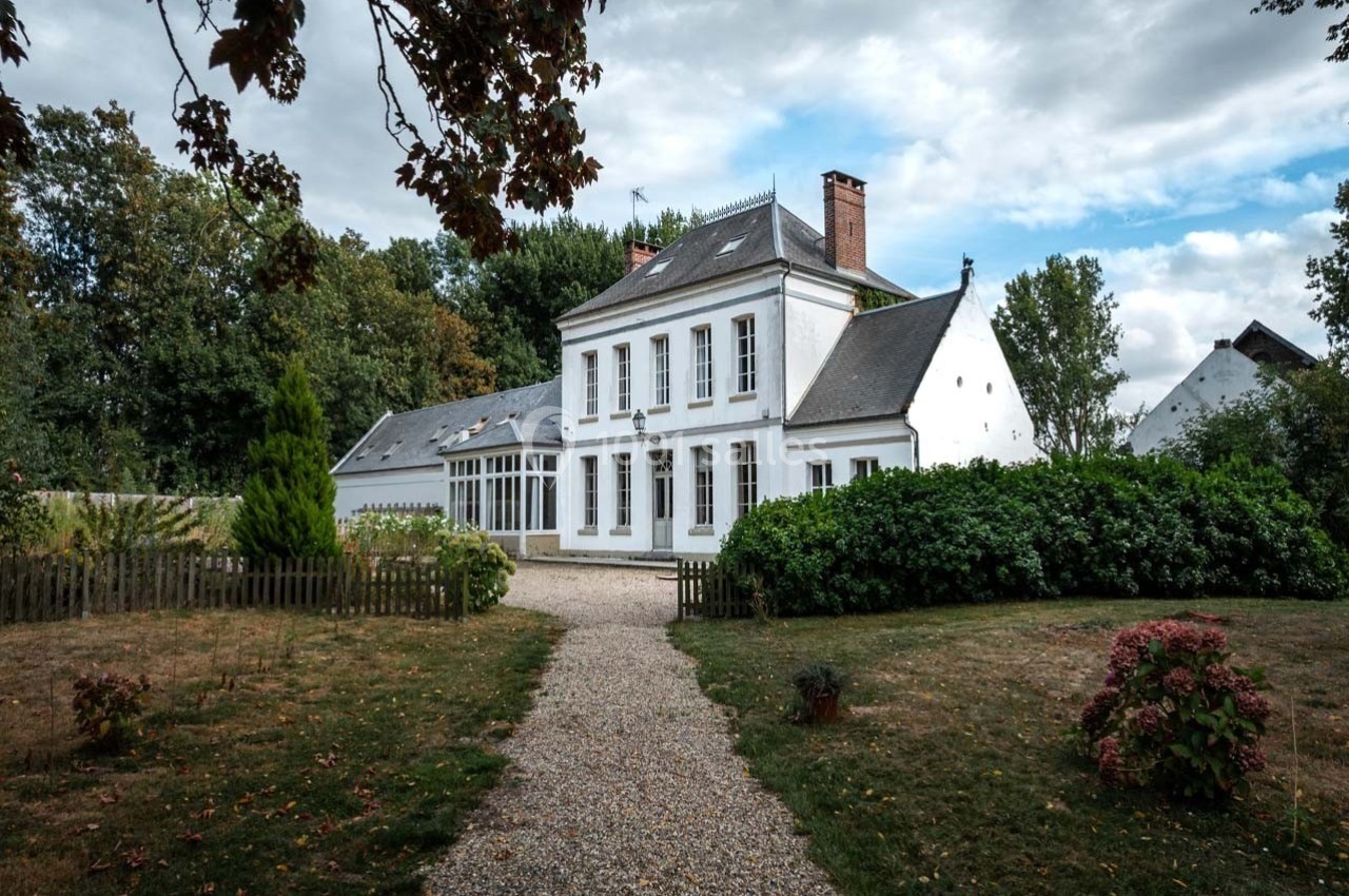 Grande maison blanche à deux étages avec jardin, allée gravillonnée et arbres en arrière-plan sous un ciel nuageux.