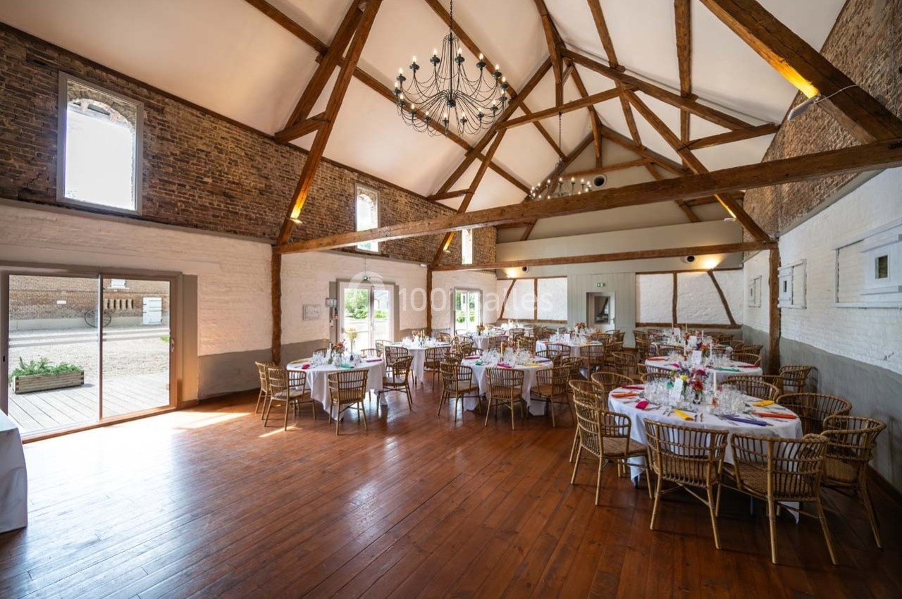 Salle de réception lumineuse avec poutres apparentes, tables rondes dressées et parquet en bois.