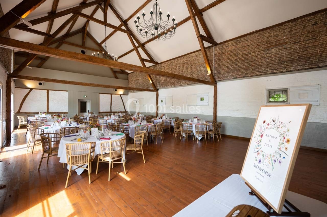 Salle de réception lumineuse avec poutres apparentes, tables dressées et panneau de bienvenue floral au premier plan.