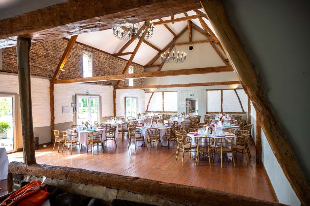 Salle de réception lumineuse avec poutres apparentes, tables rondes dressées et chaises en bois.