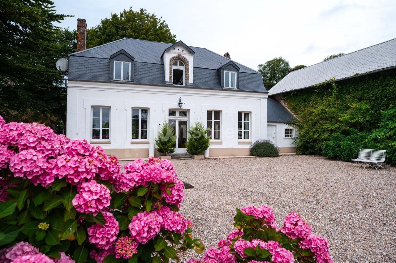 Façade d'une maison blanche avec toit en ardoise, devant une cour gravillonnée et des hortensias roses au premier plan.