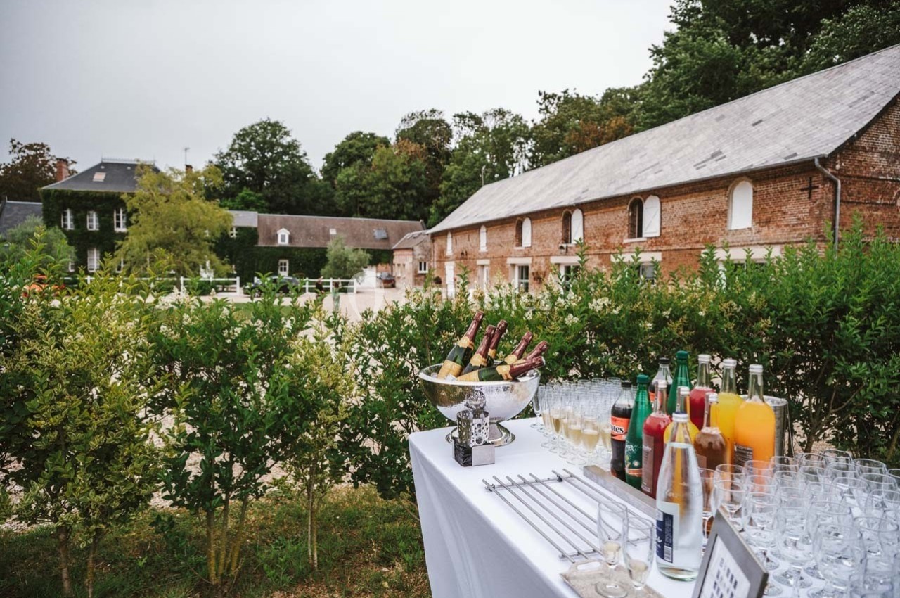 Table dressée avec boissons et verres devant un bâtiment en briques entouré de verdure et d'une cour gravillonnée.