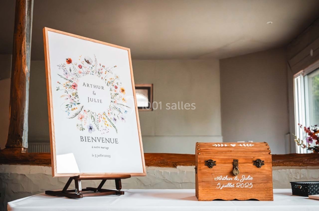 Panneau de bienvenue à un mariage avec des fleurs illustrées, posé sur un chevalet, à côté d'une boîte en bois gravée.