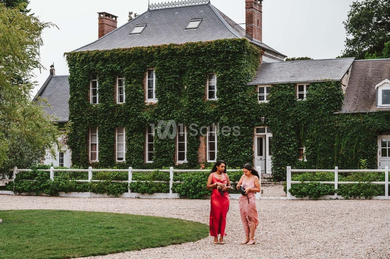 Deux femmes en robes élégantes marchent sur une allée gravillonnée devant une grande maison couverte de lierre.