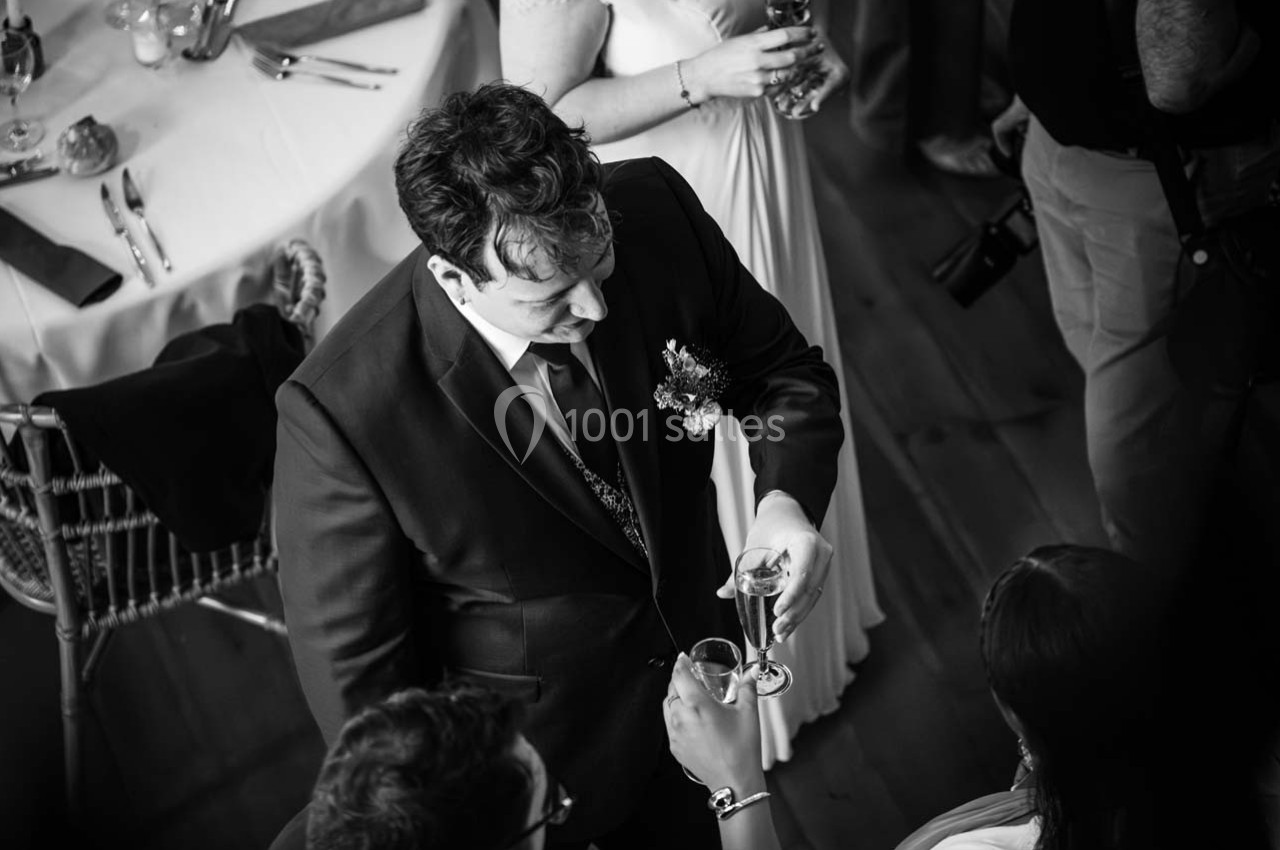 Un homme en costume sombre trinque avec des invités lors d'un événement, vu en noir et blanc.