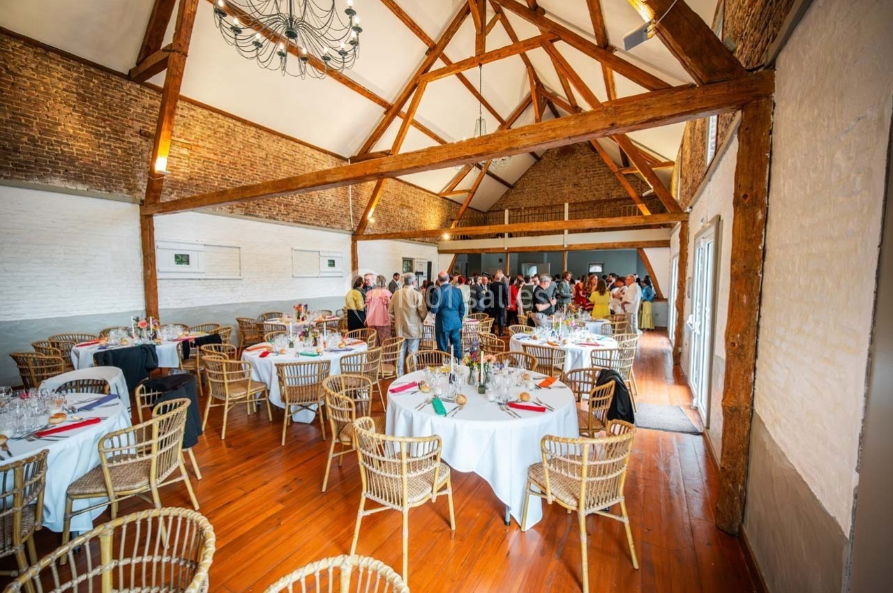 Salle de réception avec charpente en bois, tables dressées et invités rassemblés au fond de la pièce.