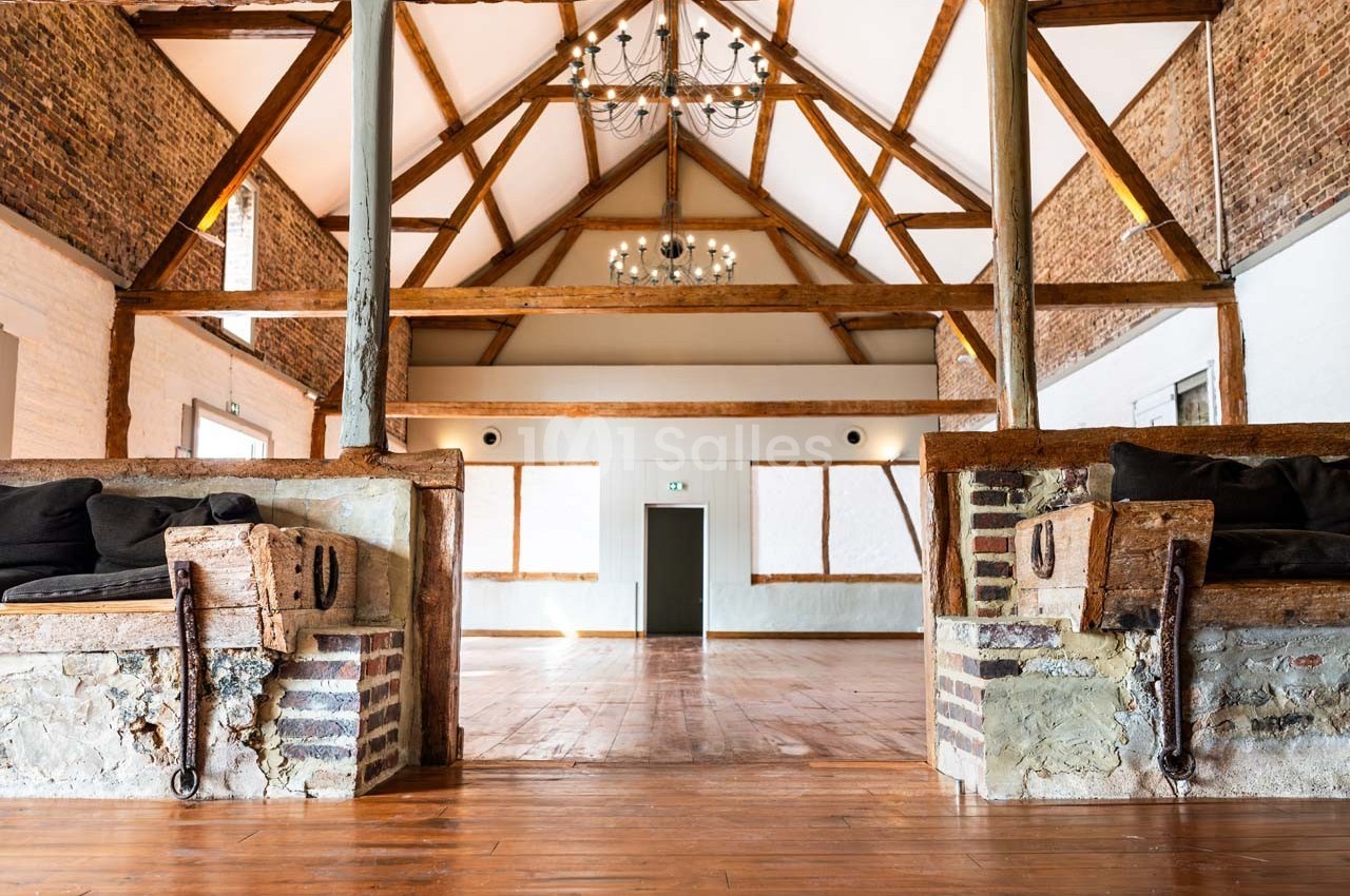 Salle spacieuse avec poutres apparentes, murs en briques et parquet en bois, éclairée par des lustres suspendus.