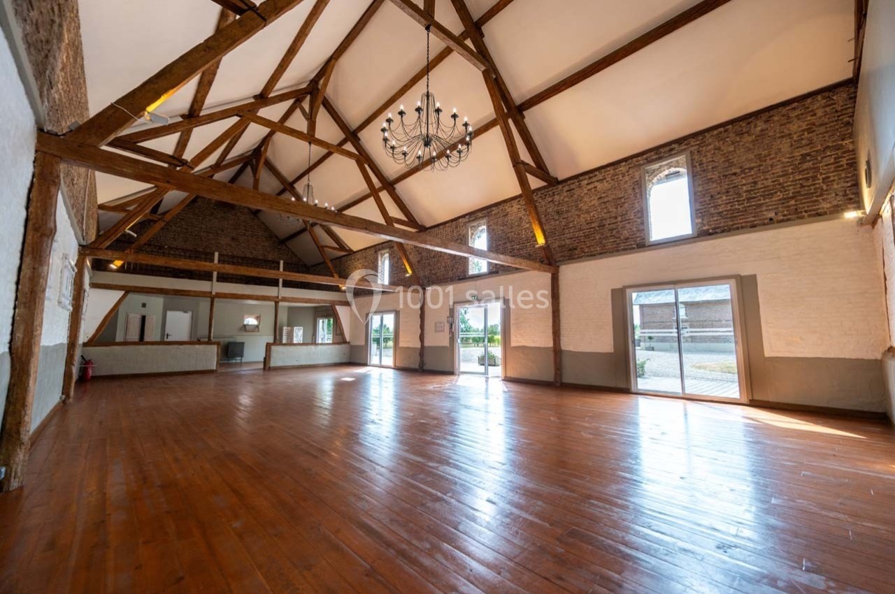 Grande salle lumineuse avec parquet en bois, poutres apparentes, murs en briques et grandes ouvertures vitrées.
