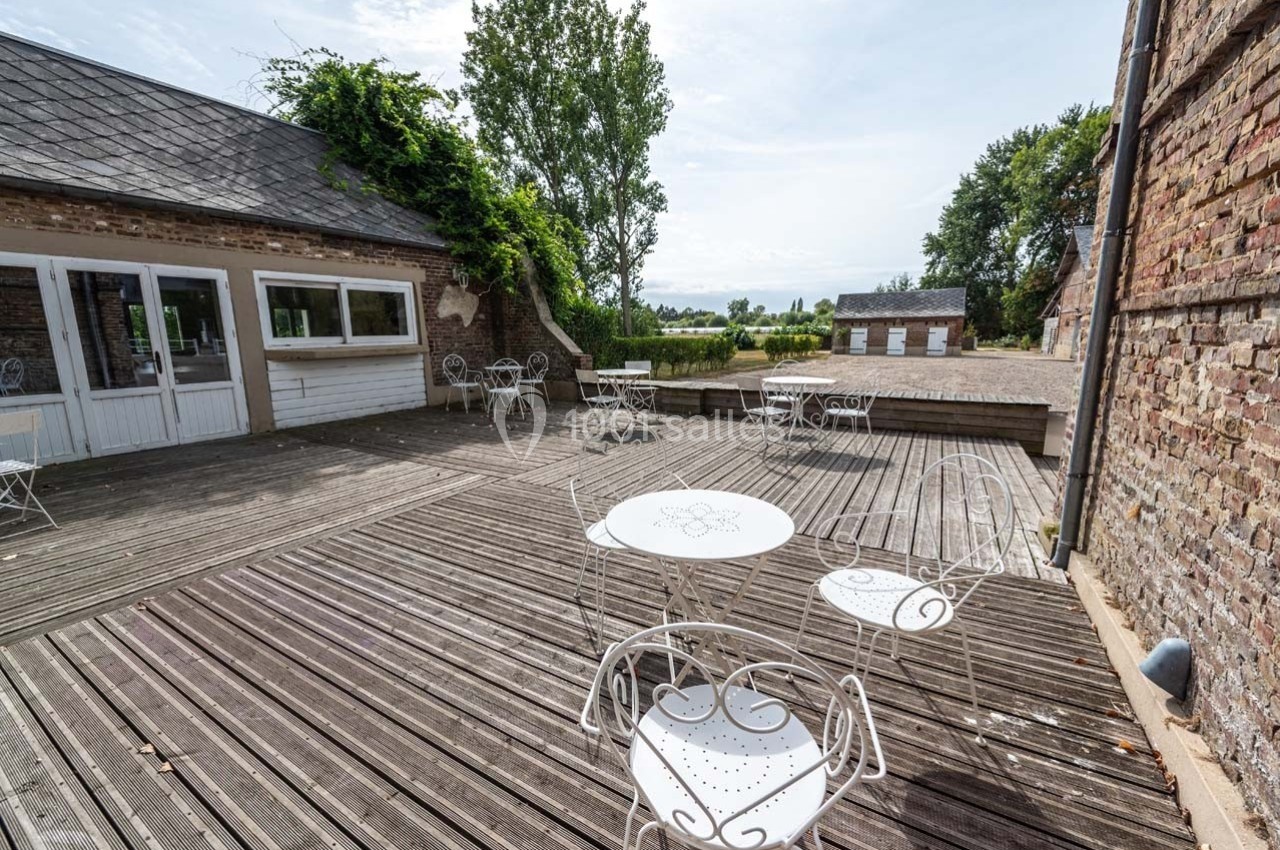 Terrasse en bois avec tables et chaises blanches, entourée de bâtiments en briques et d'arbres, vue sur la campagne.