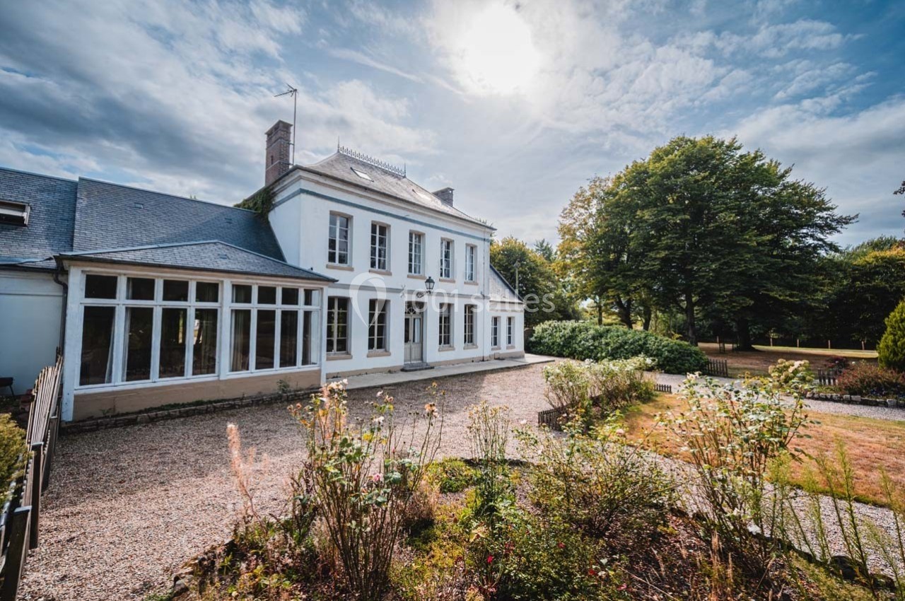 Grande maison blanche avec véranda, entourée d'un jardin fleuri et d'arbres, sous un ciel partiellement nuageux.