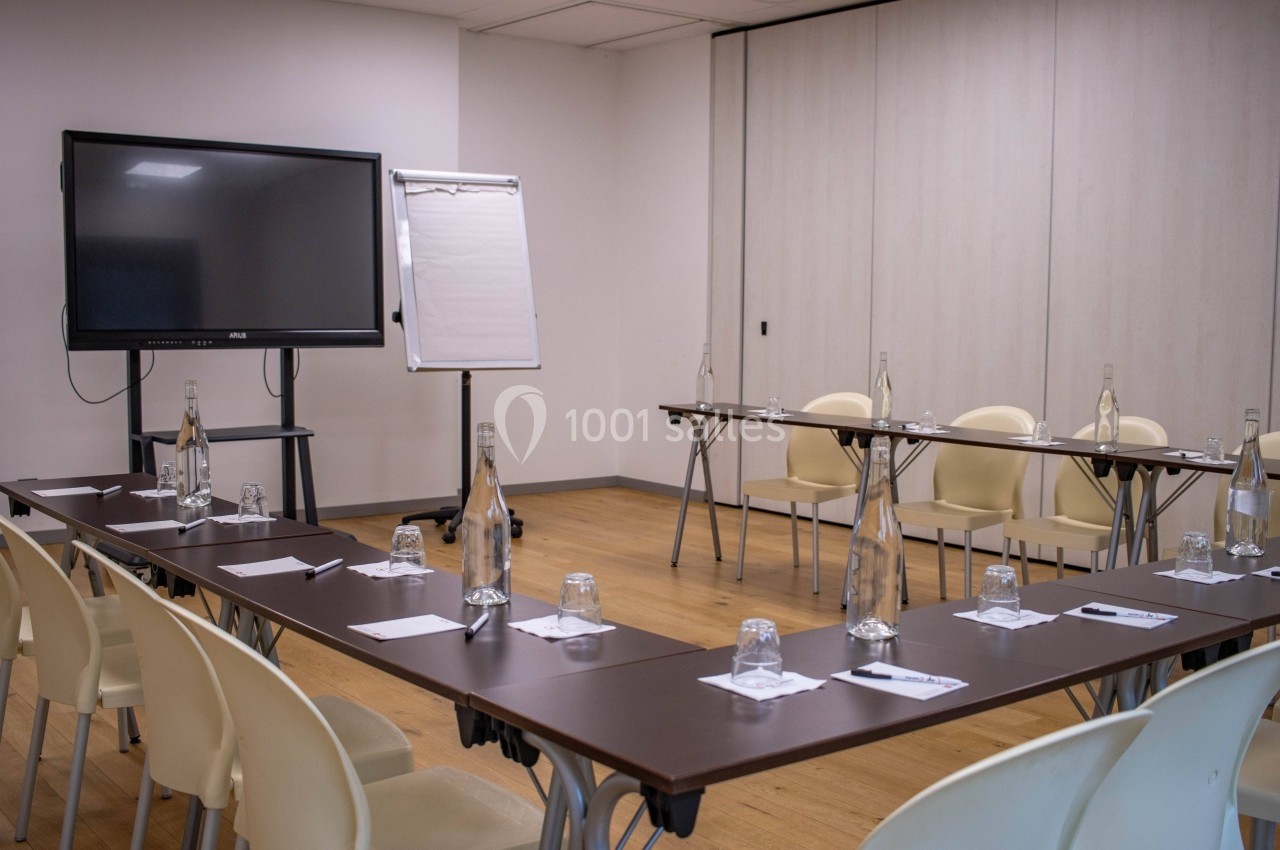 Salle de réunion avec tables disposées en U, équipée d'un écran, d'un paperboard, de bouteilles d'eau et de verres.