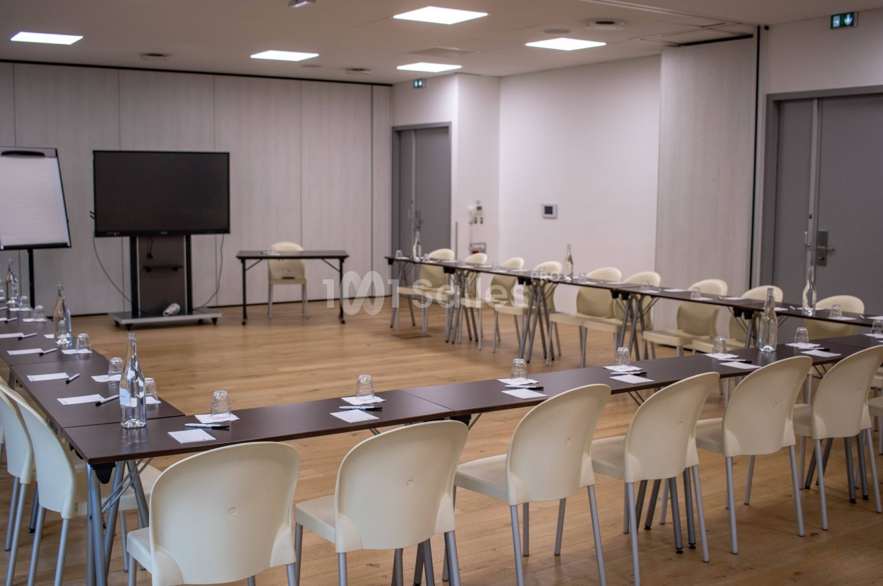 Salle de réunion lumineuse avec des tables disposées en U, des chaises, un écran et un tableau de présentation.