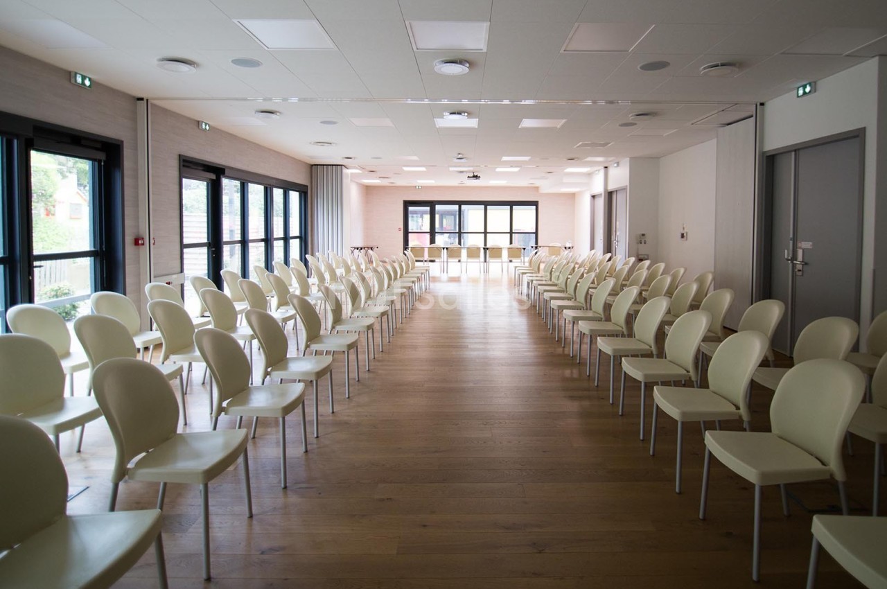 Salle de conférence lumineuse avec rangées de chaises blanches alignées sur un sol en bois clair.