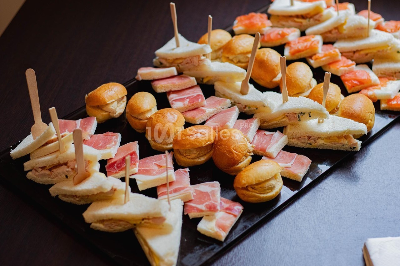 Plateau de mini-sandwichs variés avec pain blanc, jambon, saumon et brochettes en bois, disposé sur une table sombre.