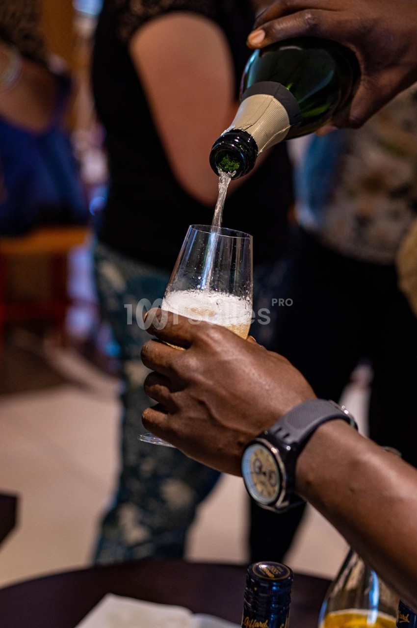 Une main verse du champagne dans un verre tenu par une autre personne, avec des silhouettes floues en arrière-plan.