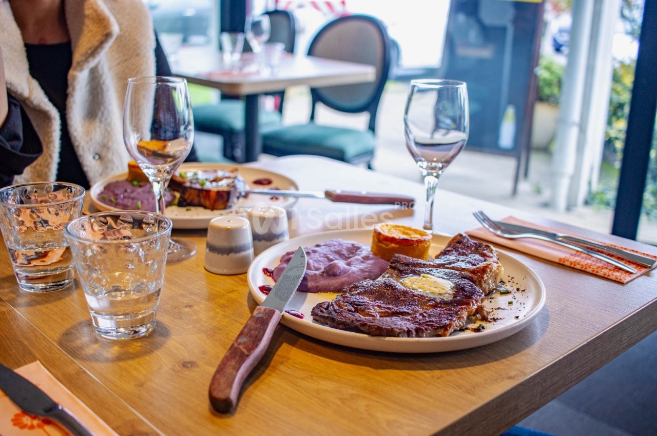 Assiette de viande grillée avec purée violette et accompagnements sur une table de restaurant, verres et couverts disposés.