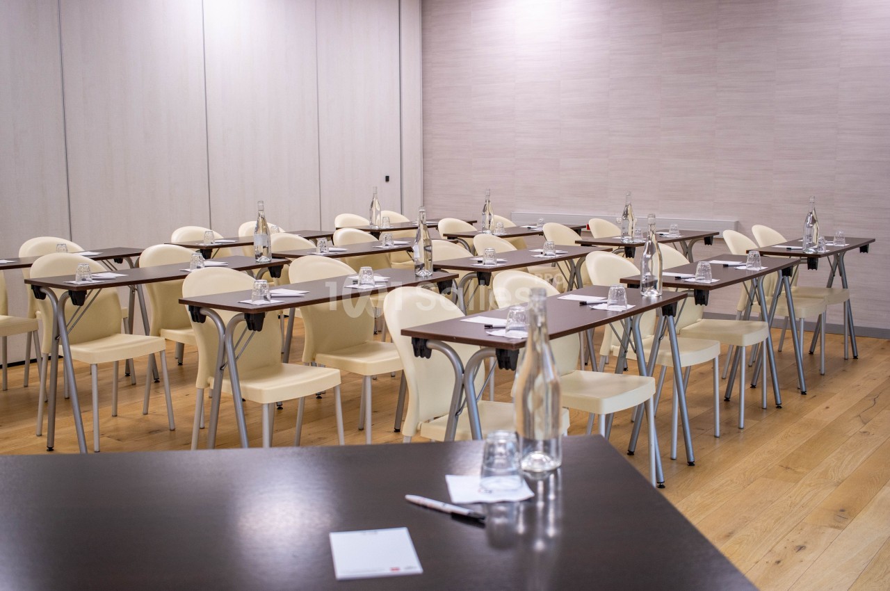 Salle de réunion avec des tables alignées, chaises beiges, bouteilles d'eau et blocs-notes sur un sol en bois.